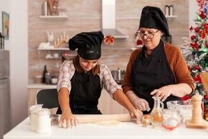 Child making gingerbread homemade dessert with grandmother celebrating christmas holiday in xmas decorated kitchen. Grandchild preparing cookies dough using baking rolling pin enjoying winter season photo