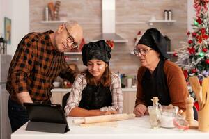 Family watching online cooking lesson on tablet standing at table in xmas decorated kitchen. Grandchild enjoying christmas season preparing gingerbread cookies dessert celebrating x-mas holiday photo