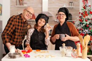 Portrait of happy family smiling while holding cookies homemade dough having tree shape celebrating christmas holiday. Grandparents enjoying winter season together with child in xmas decorated kitchen photo