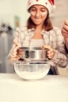 Joyfull child making cookies on christmas day sifting flour through sieve. Happy cheerful teenage girl helping senior woman preparing sweet cookies to celebrate winter holidays wearing santa hat. photo