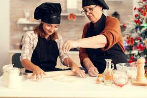 Granddaughter in apron preparing cake on christmas day with grandmother. Happy cheerful joyfull teenage girl helping senior woman preparing sweet cookies to celebrate winter holidays. photo