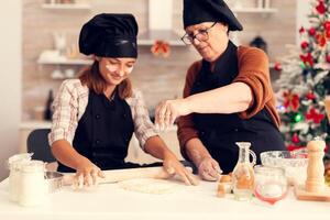 Close up of grandmother on christmas day spreading flour and making dessert with child. Happy cheerful joyfull teenage girl helping senior woman preparing sweet cookies to celebrate winter holidays. photo