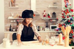 Kid on christmas day making tasty dessert using dough wearing apron. Cheerful happy cute girl while prepearing delicious cookies for christmas celebration in kitchen with christmas tree in the background. photo