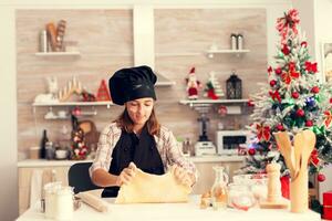 Joyful child playing with dough on christmas day wearing apron .Cheerful happy cute girl while prepearing delicious cookies for christmas celebration in kitchen with christmas tree in the background. photo