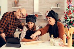 Grandparents and niece searching recipe on christmas wearing apron using tablet. Happy cheerful joyfull teenage girl helping senior woman preparing sweet cookies to celebrate winter holidays. photo