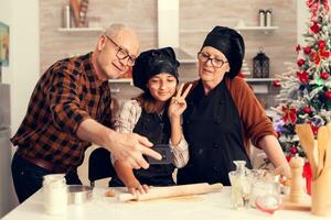 Grandparent and child taking selfie on christmas day wearingapron and bonette. Happy cheerful joyfull teenage girl helping senior woman preparing sweet cookies to celebrate winter holidays. photo