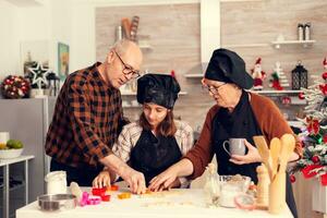 Grandparents helping niece with dessert on christmas day using shape cutter for dogh. Happy cheerful joyfull teenage girl helping senior woman preparing sweet cookies to celebrate winter holidays. photo