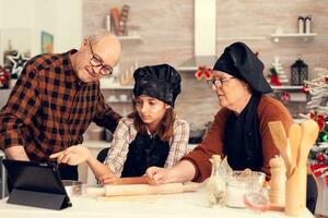 Niece and grandparents following online recipe for christmas dessert .Happy cheerful joyfull teenage girl helping senior woman preparing sweet cookies to celebrate winter holidays. photo