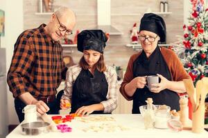 Granddaughter on christmas day having assistance on making dessert from grandparents. Happy cheerful joyfull teenage girl helping senior woman preparing sweet cookies to celebrate winter holidays. photo