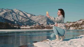 une femme touristique est assis sur le rivière banque dans une montagneux zone et prend une selfie en utilisant une téléphone intelligent. video