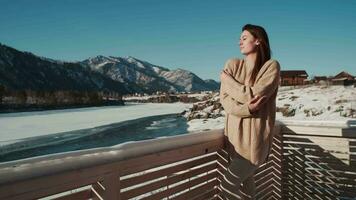 ein Frau im ein Jacke steht auf das Balkon und entspannt sich gegen das Hintergrund von Berge im Winter. video