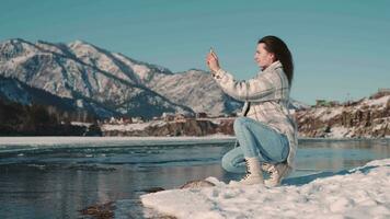 een vrouw toerist blogger zit Aan de rivier- bank in een bergachtig Oppervlakte en duurt een foto gebruik makend van een smartphone. video