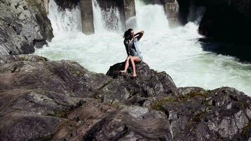 lindo menina posando sentado em a pedras. atrás dela cai cascata video