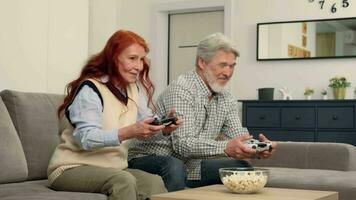 Cheerful senior couple 50-60 years old play video game at home sitting on sofa pushing each other.