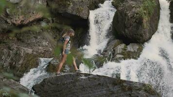 une femelle touristique grimpe une Roche et soulève le sien mains en haut dans de face de une grand cascade. content fille jouit Succès video