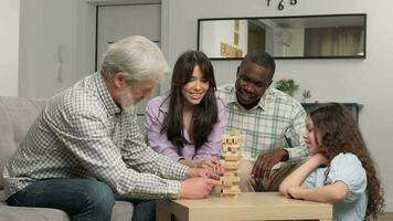 ein multiethnisch Familie von anders Alter Generationen ist spielen ein Tafel Spiel beim Zuhause entfernen hölzern Blöcke von ein Turm. video