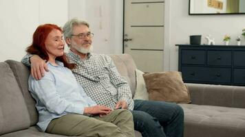 Happy senior couple aged 60-70 watching news on TV while sitting on the couch at home and talking. video