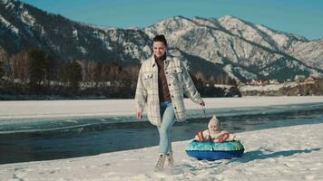 A happy mother rolls her daughter on a sled near the river in winter. video