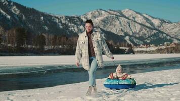 A happy mother rolls her daughter on a sled near the river in winter. video