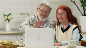 Happy senior couple in their 60s and 70s make a video call while sitting at a table in the kitchen using a laptop.