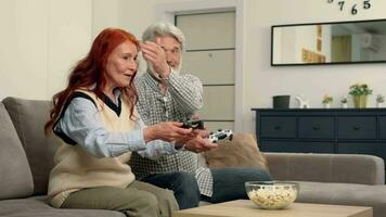 Cheerful senior couple 50-60 years old playing a video game at home sitting on the couch. Grandpa covers grandma's eyes.