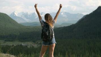 ragazza viaggiatore con un' zaino nel pantaloncini sta a il picco di il montagna e solleva mani su. obbiettivo realizzazione video