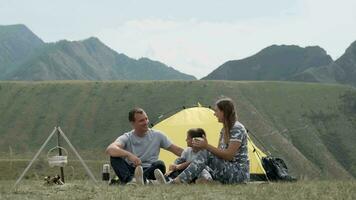 un contento familia con un niño es descansando juntos en frente de un tienda en el montañas y Bebiendo té desde un termo. video