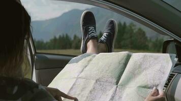 le turisque fille regards à une carte tandis que séance dans une voiture et collage le sien jambes en dehors le fenêtre contre le toile de fond de le montagnes. voiture à l'intérieur vue video