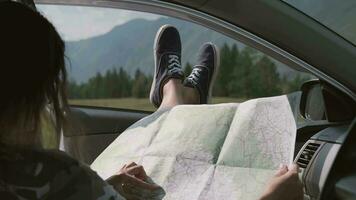 le turisque fille regards à une carte tandis que séance dans une voiture et collage le sien jambes en dehors le fenêtre contre le toile de fond de le montagnes. video