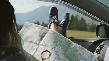 ein turisk Mädchen sieht aus beim ein Karte mit ein Lupe während Sitzung im ein Auto und kleben seine Beine aus das Fenster gegen das Hintergrund von das Berge. video