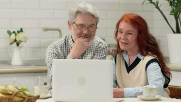 vrolijk senior paar in hun Jaren 60 en jaren 70 maken een video telefoontje terwijl zittend Bij een tafel in de keuken gebruik makend van een laptop.