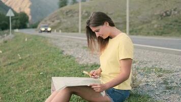 hermosa niña turista mira a un mapa de el carreteras mientras sentado en el autopista en verano soleado clima. lado ver video