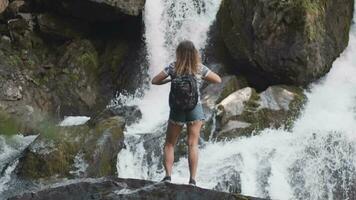 weiblich Tourist mit ein Rucksack erhöht seine Hände oben Gegenteil ein groß Wasserfall Stehen auf das Kante von ein Cliff. glücklich Mädchen genießt Erfolg. schleppend Bewegung video