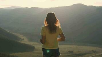 een jong vrouw komt naar de rand van een klif en verhoogt haar handen omhoog in voorkant van de hoog rotsachtig bergen gedurende zonsondergang. gelukkig meisje geniet succes video