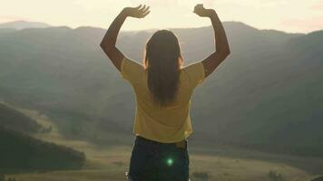 uma jovem mulher vem para a Beira do uma penhasco e levanta dela mãos acima dentro frente do a Alto rochoso montanhas durante pôr do sol. feliz menina goza sucesso. sucesso conceito video