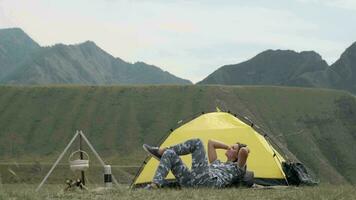 Girl tourist with gray clothes relaxes lying in front of the tent on a background of mountains in sunny weather video