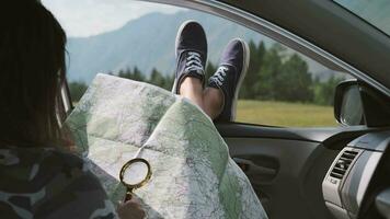 Girl looks at a map using a magnifier while sitting in a car and sticking his legs out the window against the backdrop of the mountains video