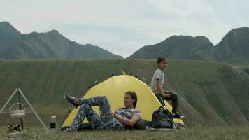 Mama und Sohn sind ruhen auf ein Campingplatz im Vorderseite von das Zelt. das Junge Blätter das Zelt nach Schlafen und zieht seine Hände hoch. video