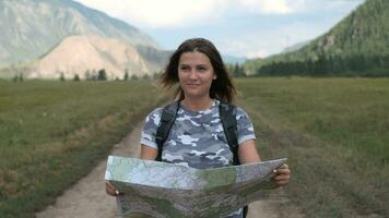 giovane donna turista con un' zaino va su il strada con un' carta geografica nel mani su un' sfondo di montagne. viaggio concetto video