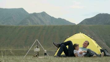 Father and son are resting lying in front of a tent and a bone in sunny weather on a background of mountains video