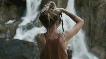 Young paparazzi girl photographs a beautiful large waterfall standing on the stones in the jungle video
