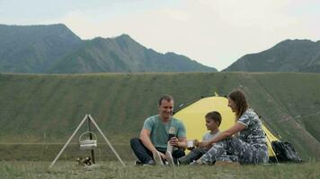 glücklich jung Familie mit ein Kind ruhen zusammen im Vorderseite von ein Zelt im das Berge und Trinken Tee von ein Thermosflasche. video