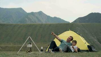 Happy father and son relax together and rejoice in front of the tent against the backdrop of the mountains. video