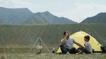 Happy Father and son are sitting near the tent and bonfire and laughing. Camping video