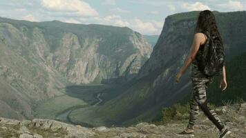 jovem menina turista com grandes cabelo com uma mochila abordagens a pico do a montanha e parece frente video