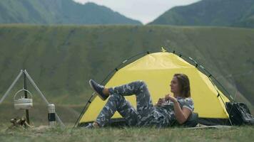 ragazza turista riposo dire bugie nel davanti di un' tenda con un' boccale di tè su un' sfondo di montagne video