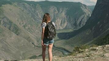 un hembra viajero con un mochila sube a el parte superior de el montaña y levanta su mano arriba con un sensación de apuesto y éxito. espalda ver video