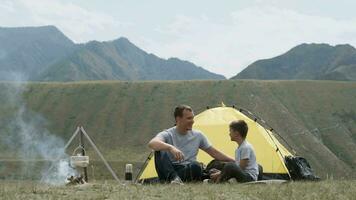 Father and son are sitting near the tent and bonfire and laughing. video