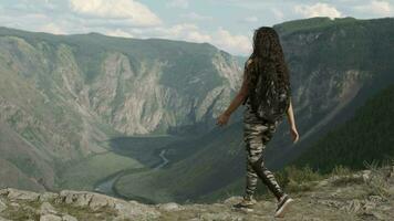 lindo menina turista com grandes cabelo com uma mochila vem para a pico do a montanha e regozija-se levantando braços acima video