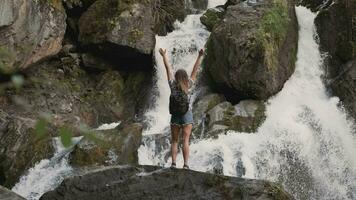 kvinna turist med en ryggsäck höjer hans händer upp motsatt en stor vattenfall stående på de kant av en klippa. Lycklig flicka åtnjuter Framgång video